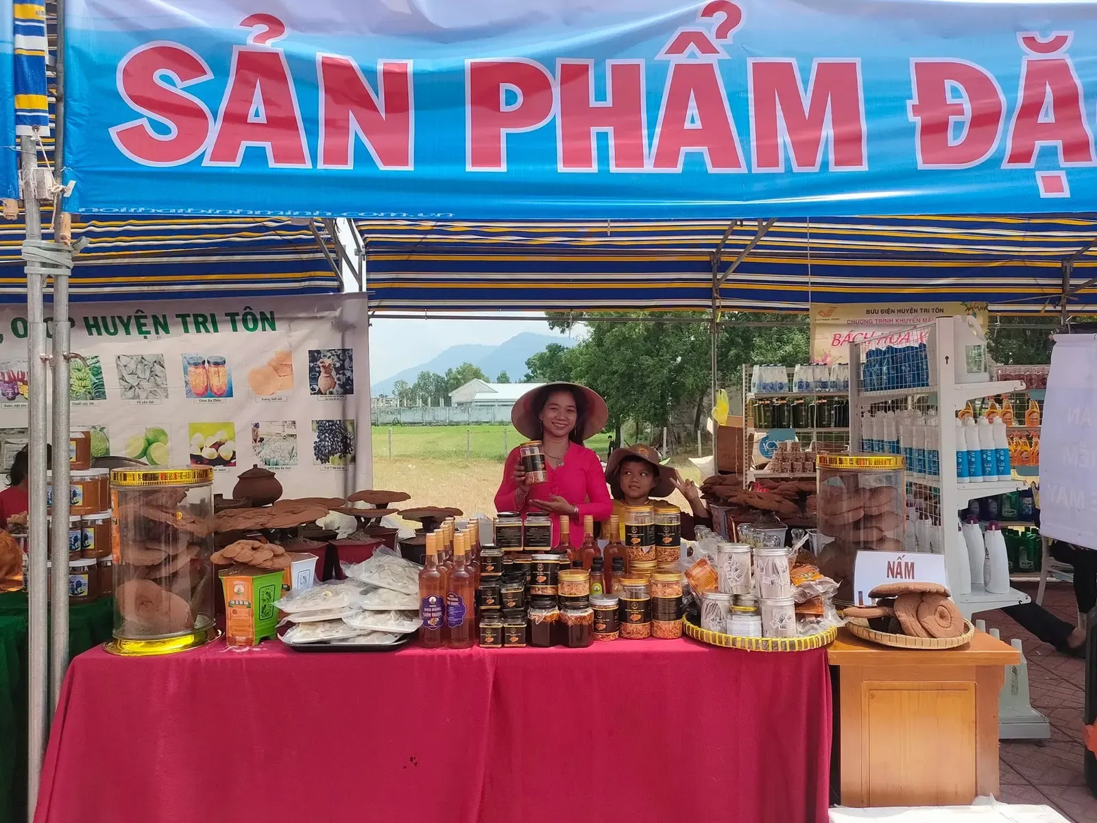 Gian hàng hội chợ dưới băng rôn sản phẩm đặc trưng: nấm linh chi, hũ đông trùng hạ thảo và các sản phẩm từ nấm bày trên bàn phủ khăn đỏ, phía sau là bảng sản phẩm OCOP huyện Tri Tôn