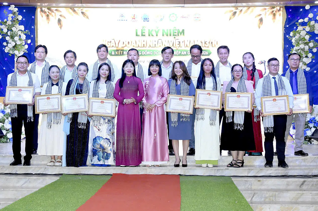 Hai hàng đại biểu quàng khăn rằn, nhiều người cầm bằng khen, đứng trên sân khấu trước phông nền Lễ kỷ niệm Ngày Doanh nhân Việt Nam 13/10; chị Châu Thị Nương mặc áo dài hoa sen xanh đứng thứ tư từ trái hàng trước