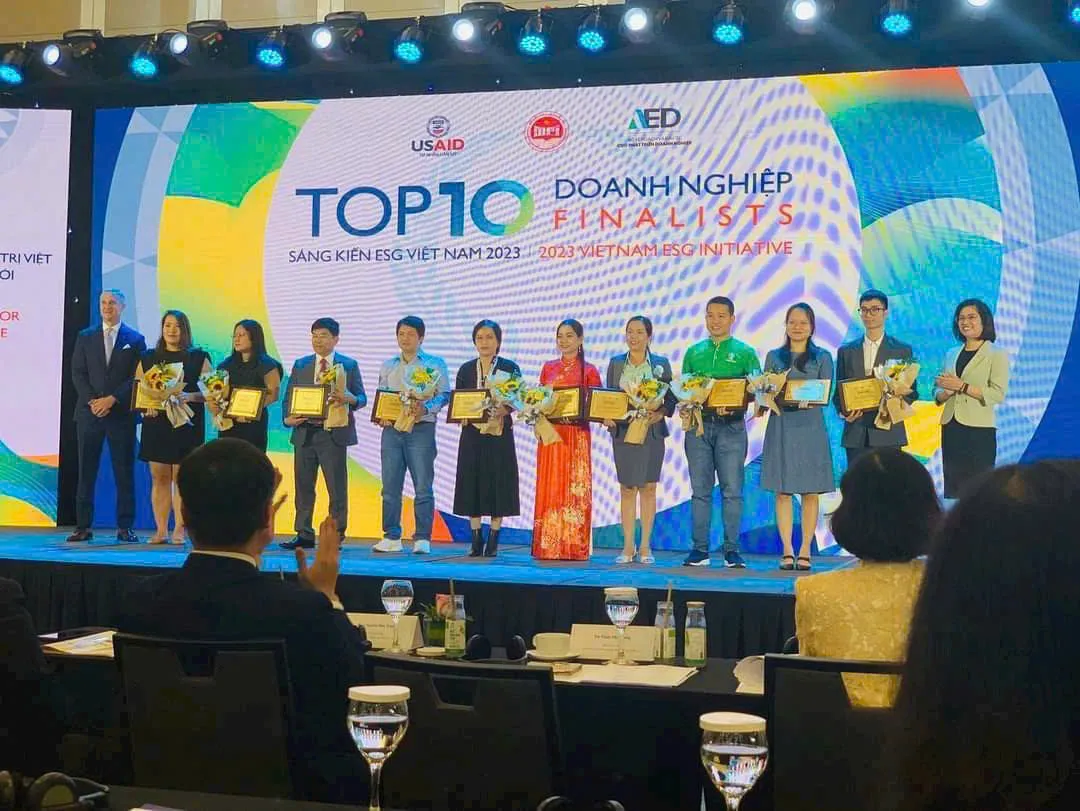 Mười đại diện doanh nghiệp đứng thành hàng trên sân khấu, cầm hoa và chứng nhận, chị Châu Thị Nương mặc áo dài đỏ ở giữa; màn hình phía sau ghi Top 10 doanh nghiệp Finalists, Sáng kiến ESG Việt Nam 2023, cùng logo USAID