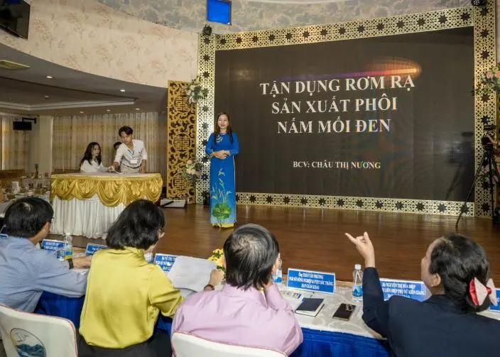 Người thuyết trình đứng trên sân khấu trước màn hình ghi Tận dụng rơm rạ sản xuất phôi nấm mối đen, BCV: Châu Thị Nương; hội đồng giám khảo ngồi phía trước