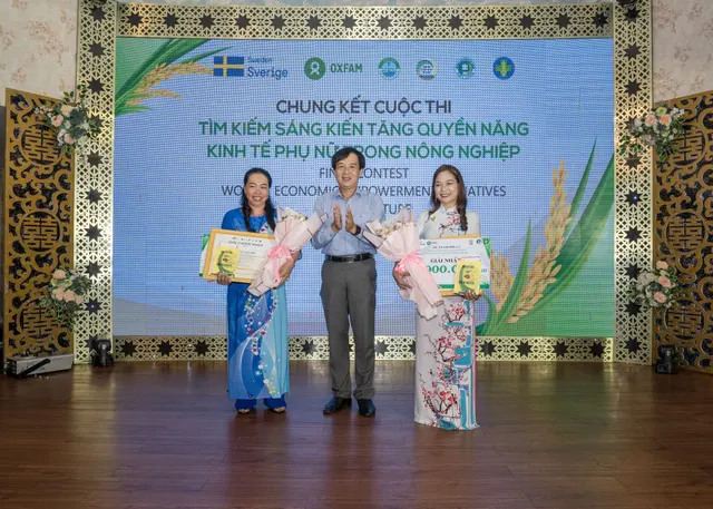 Trên sân khấu chung kết cuộc thi, hai phụ nữ mặc áo dài cầm hoa và bảng giải thưởng đứng hai bên đại diện ban tổ chức; người bên phải cầm bảng Giải Nhất