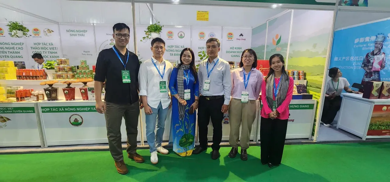 Sáu người đeo thẻ triển lãm đứng chụp ảnh chung trên thảm xanh trước dãy gian hàng, trong đó có chị Châu Thị Nương mặc áo dài xanh quàng khăn rằn; phía sau là kệ chậu nấm linh chi