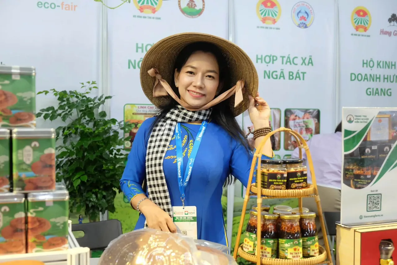 Chị Châu Thị Nương mặc áo dài xanh, quàng khăn rằn, đội nón lá, đeo thẻ triển lãm, đứng tại gian trưng bày bên giỏ mây xếp các hũ sản phẩm