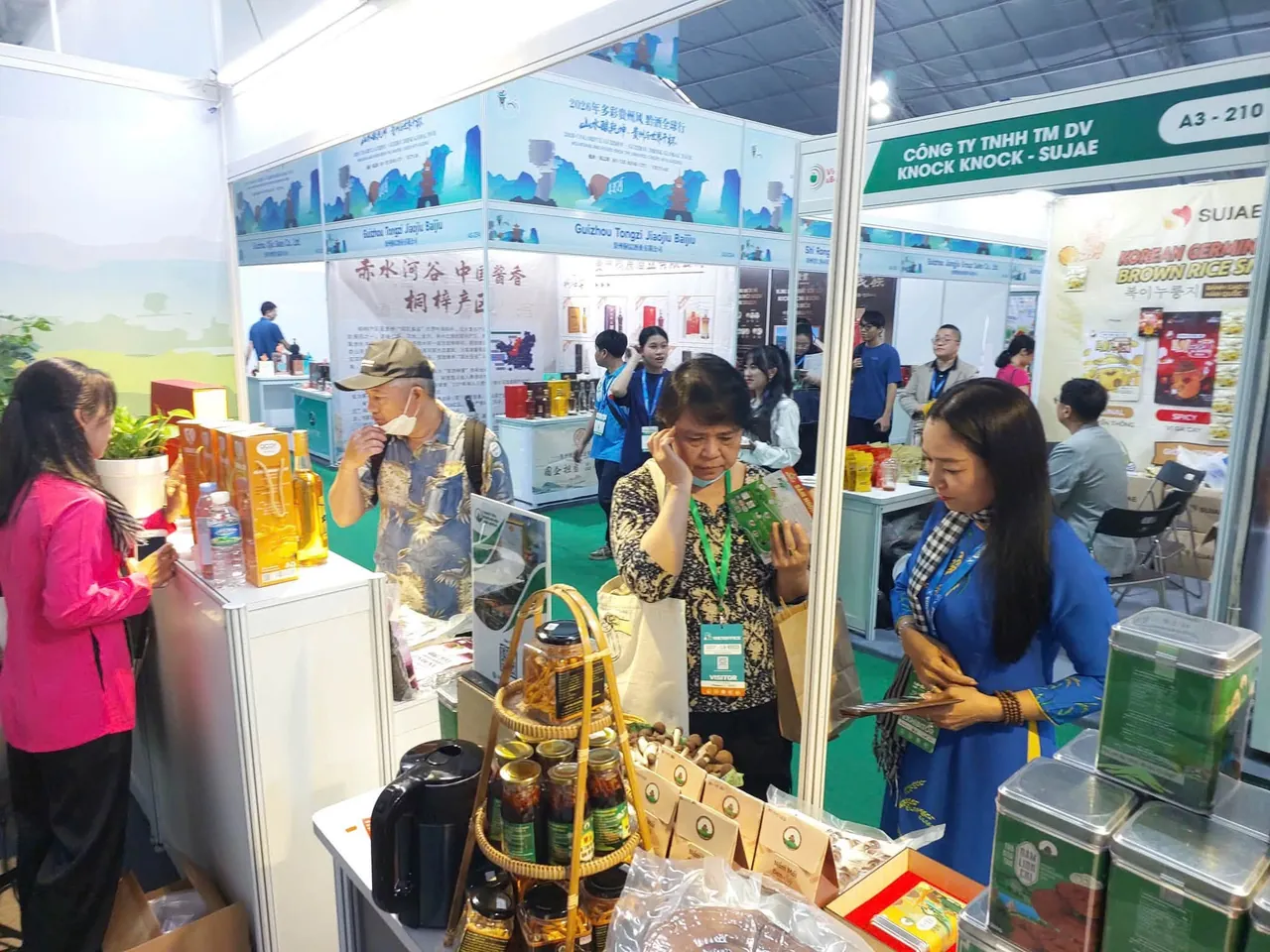 Chị Châu Thị Nương mặc áo dài xanh, quàng khăn rằn, trao đổi với khách tham quan đang xem tờ giới thiệu, bên giỏ mây xếp hũ sản phẩm và các hộp thiếc in hình nấm linh chi