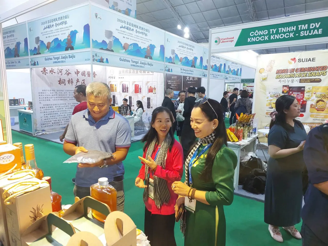 Hai phụ nữ quàng khăn rằn, một người áo đỏ và một người áo dài xanh lá, trò chuyện với một vị khách đang cầm xem gói sản phẩm; phía sau là các gian hàng khác của triển lãm