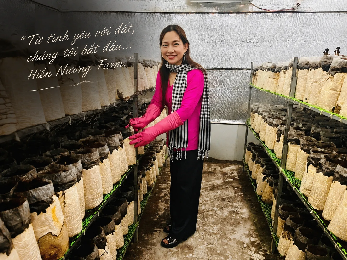 Chị Châu Thị Nương mặc áo hồng, quàng khăn rằn, đứng giữa hai dãy kệ bịch phôi trong phòng nuôi nấm. Dòng chữ viết tay trên ảnh: “Từ tình yêu với đất, chúng tôi bắt đầu... Hiền Nương Farm.”